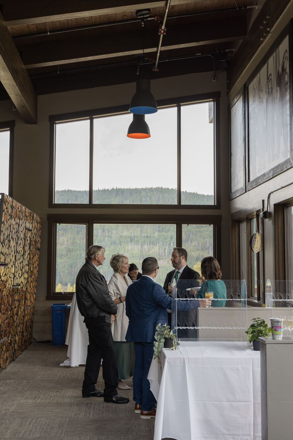 Guests mingle at an elegant indoor event venue with large windows overlooking a forested mountain landscape.