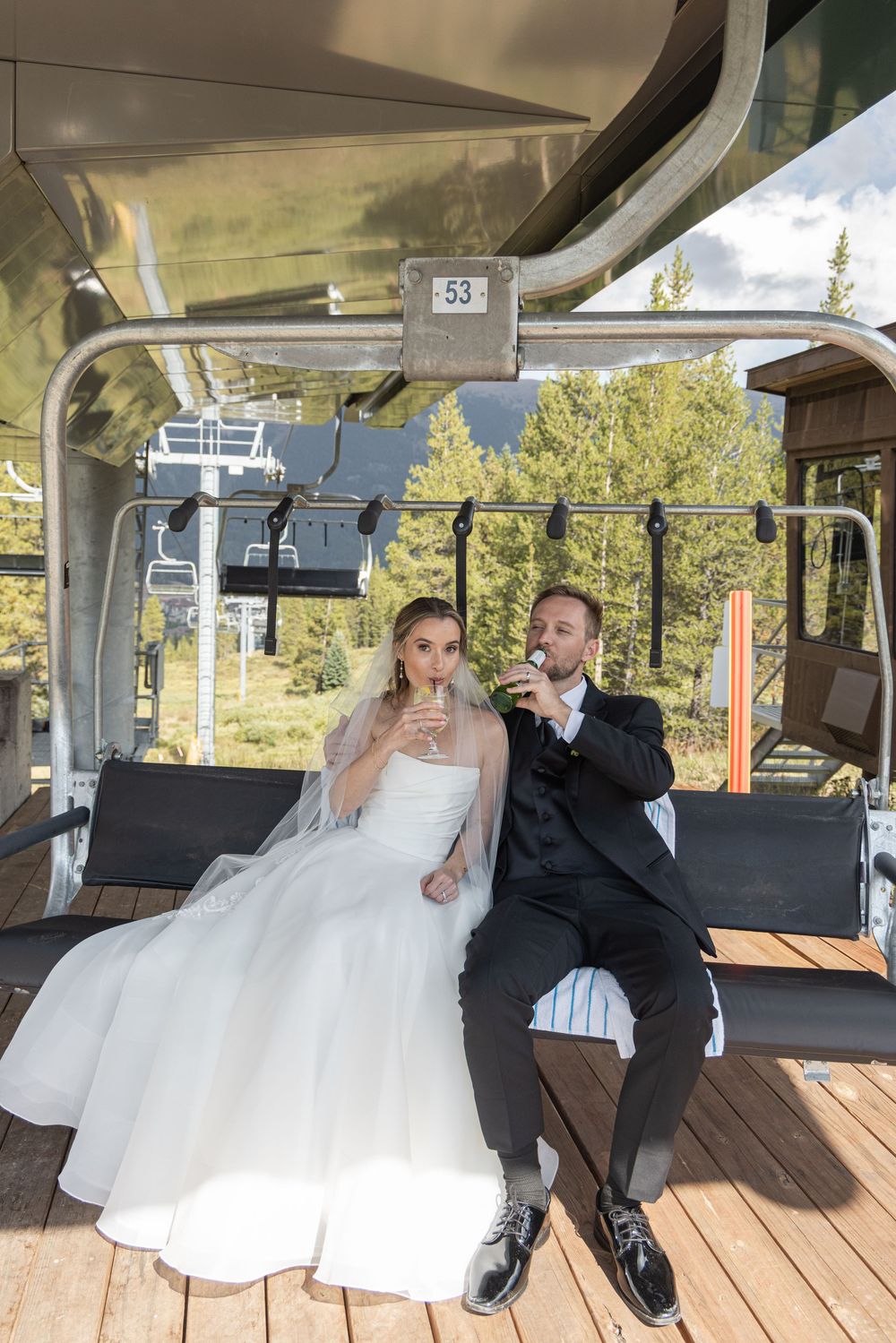 Bride and groom in wedding attire toasting champagne on ski lift gondola number 53 with forest backdrop.