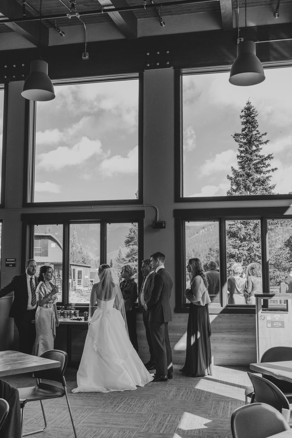 Black and white photo of a bride and groom exchanging vows indoors with large windows and a tree visible outside.