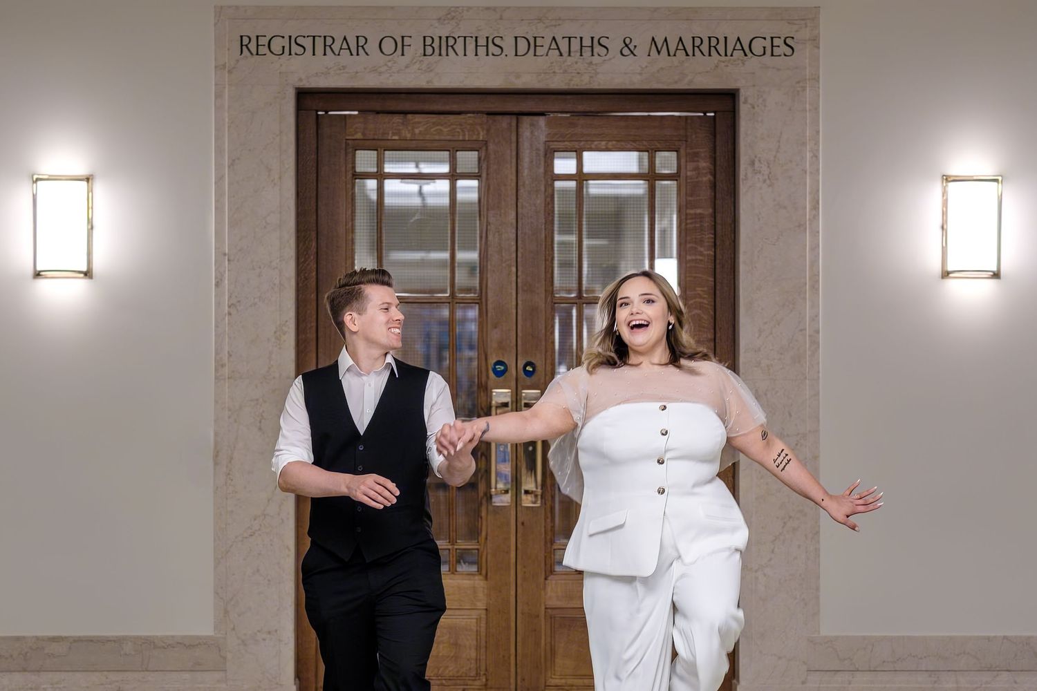 Joyful couple exits Registrar of Births Deaths and Marriages office after wedding ceremony in elegant white attire.