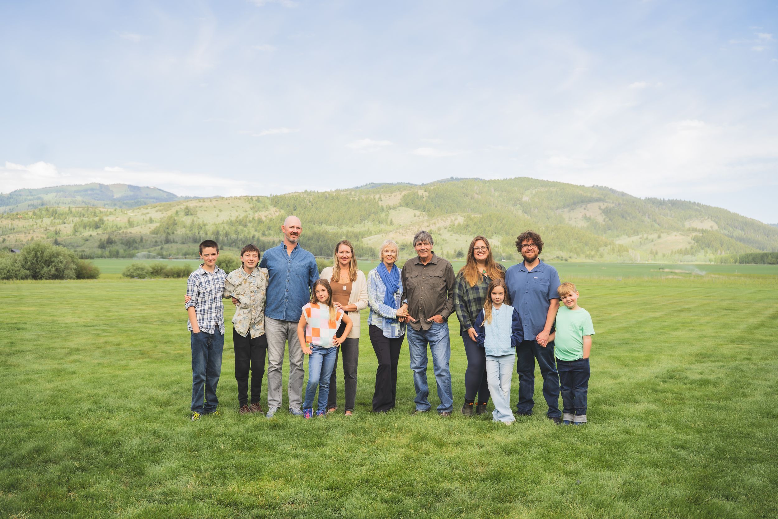 Multi-generational family portrait smiling together on a lush green field with rolling hills in the background.