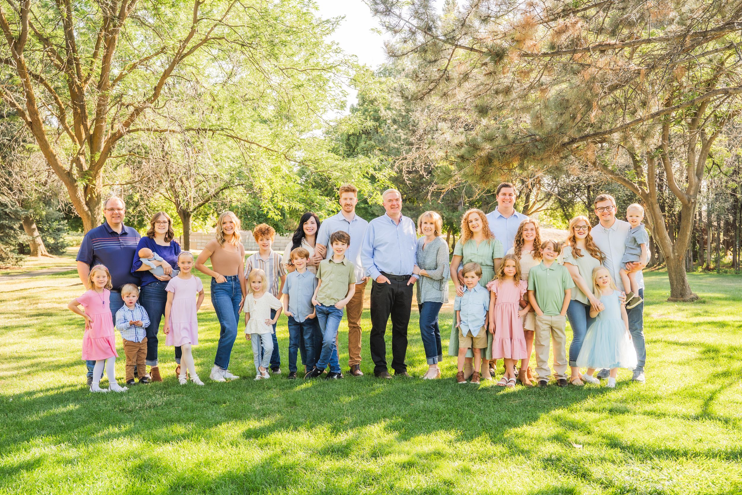 Large multigenerational family portrait outdoors on green lawn surrounded by trees on a sunny day.