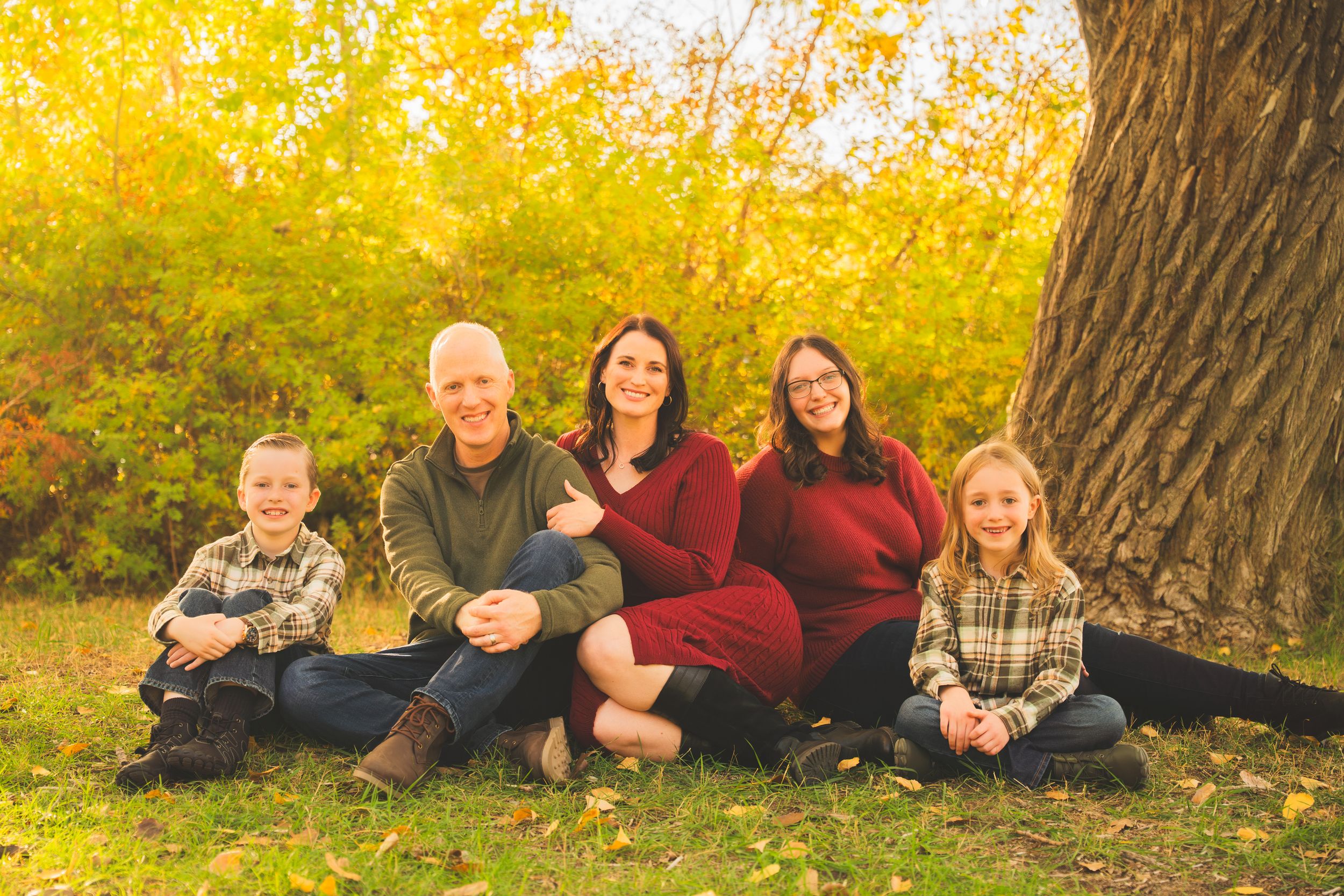Family of five smiling together outdoors in autumn, sitting near a large tree with golden fall foliage.