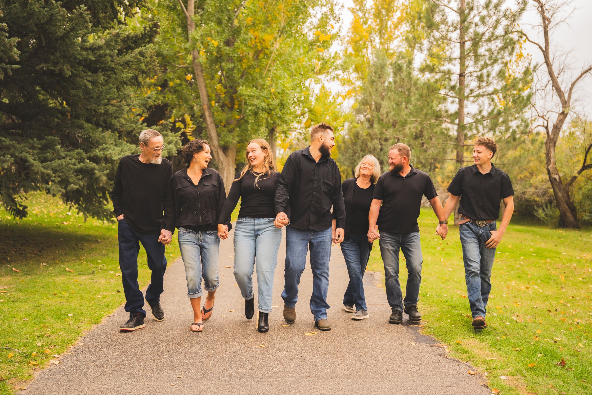 Seven people in matching black tops and jeans walk together on a park path surrounded by lush green trees.