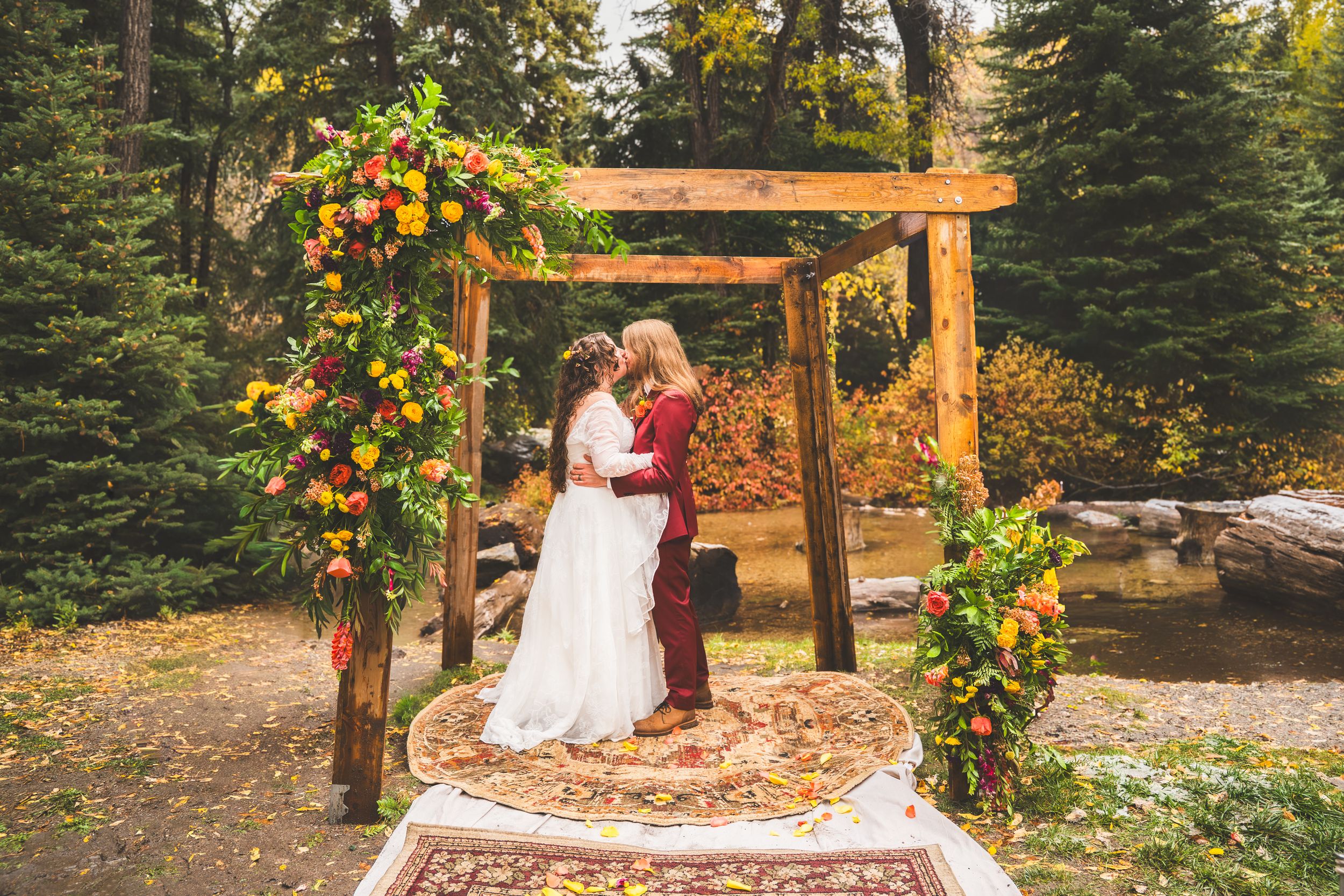 Same-sex couple shares first kiss under rustic floral arch with autumn foliage at outdoor forest wedding ceremony.