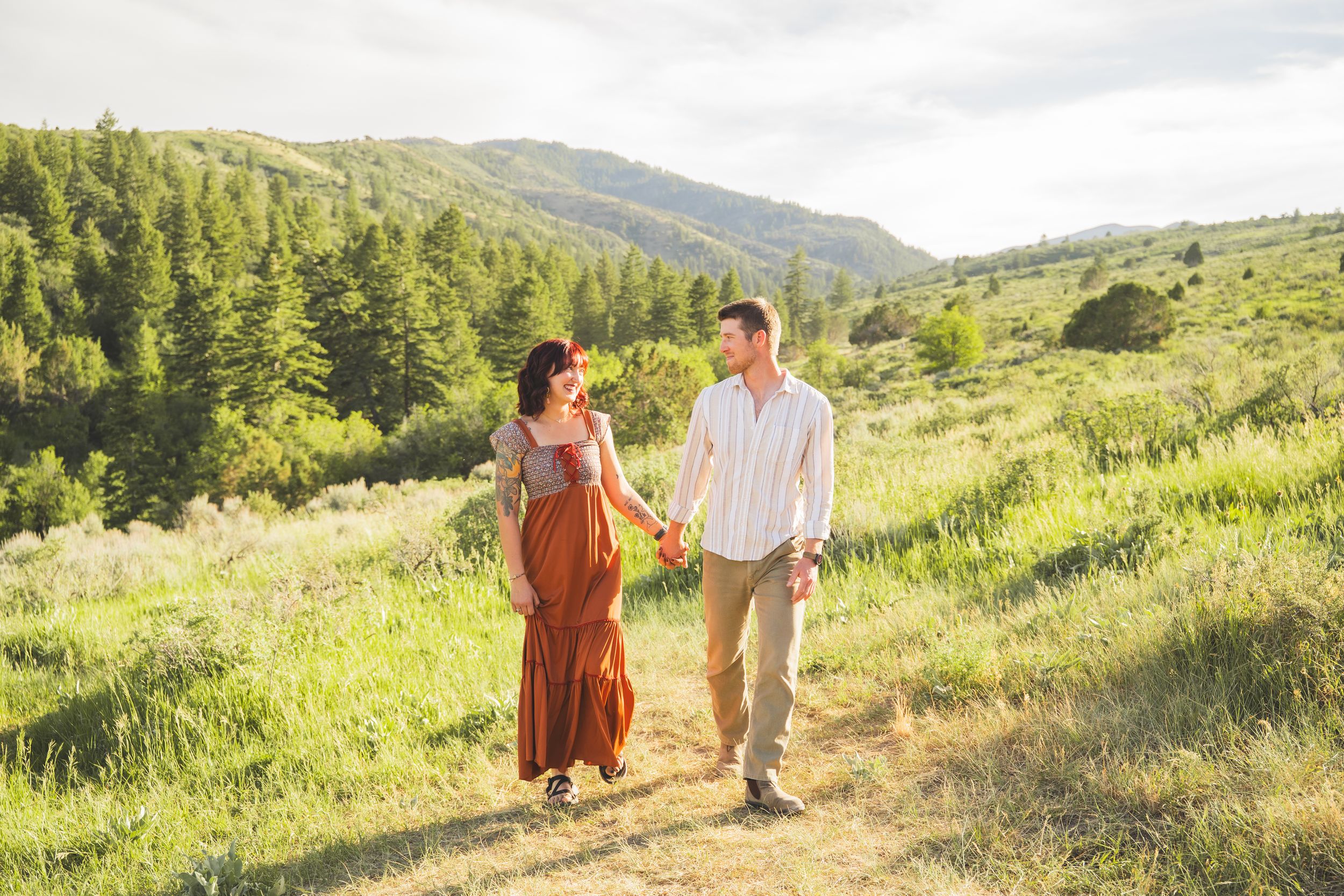 A couple walks hand-in-hand through a sunny green meadow with pine-covered hills in the background.