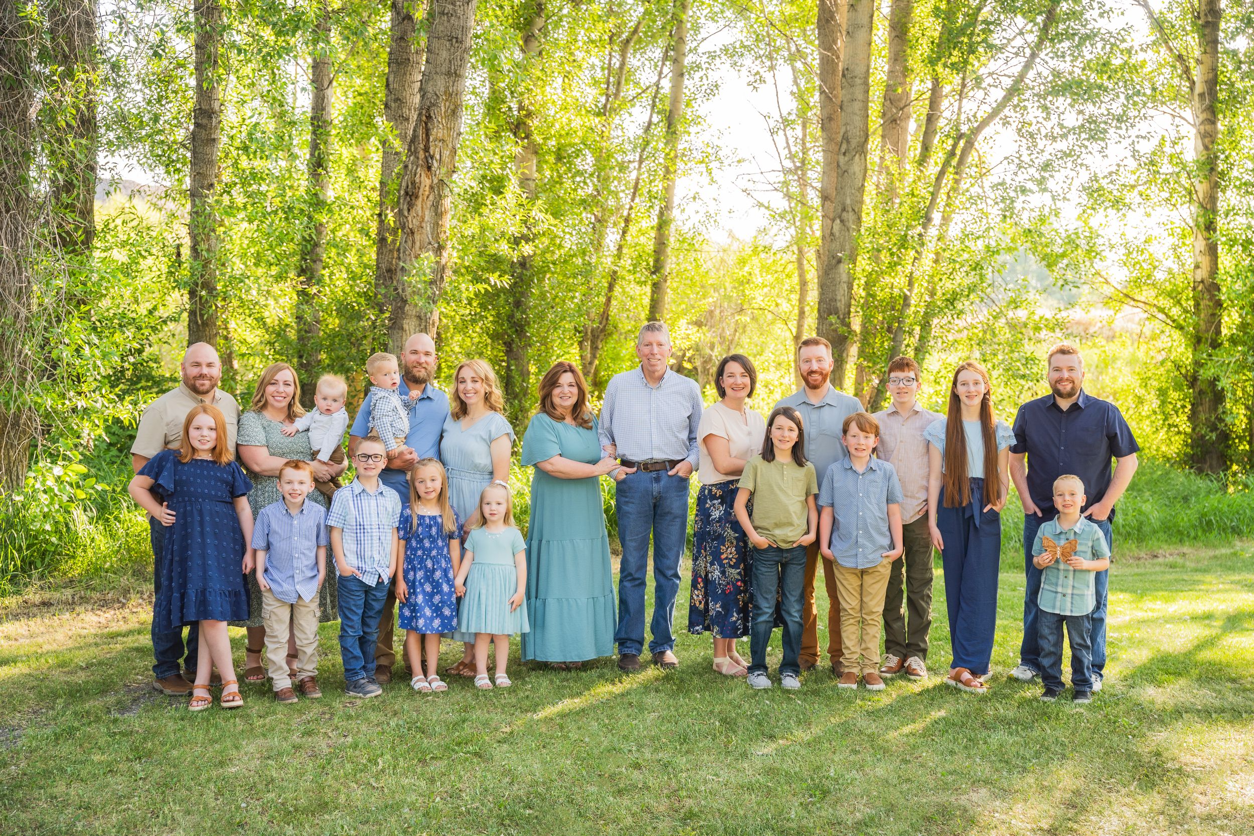 Large multi-generational family portrait outdoors on green lawn surrounded by tall trees in warm sunlight.