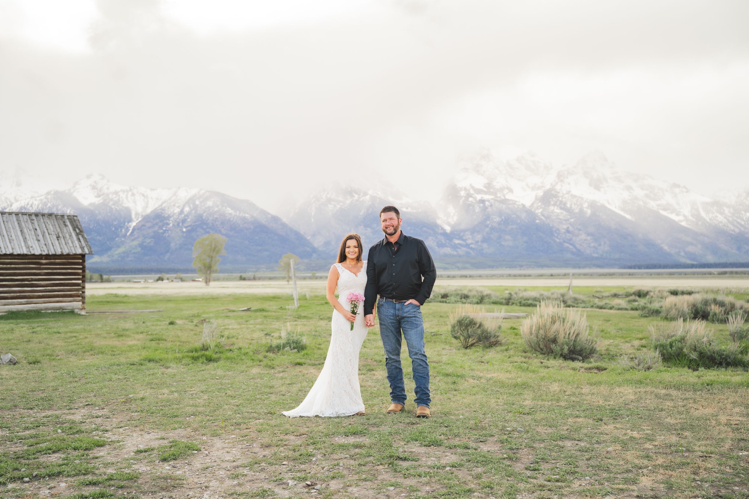Couple poses in wedding attire before snow-capped Grand Teton mountains in Wyoming with rustic barn nearby.