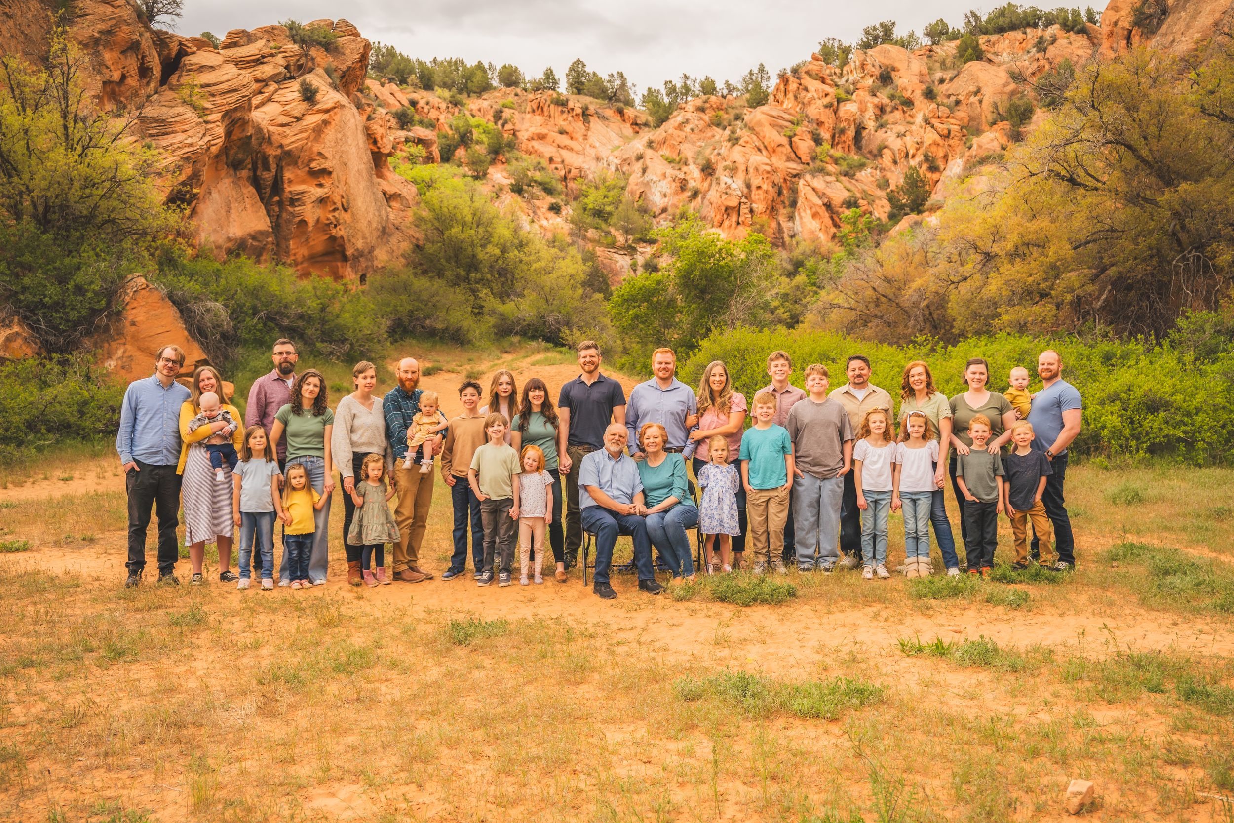 Large family group portrait outdoors with red rock formations and green trees in background on a sunny day.