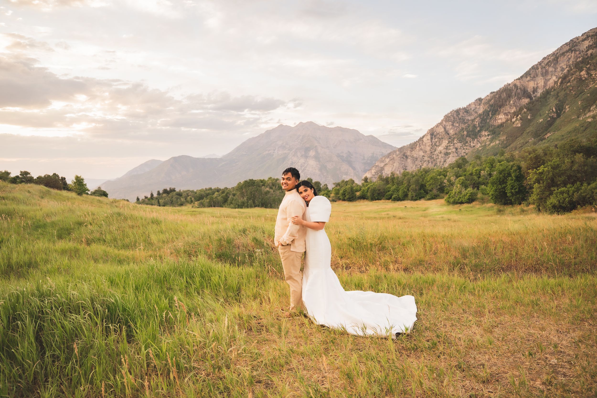 Bride and groom embracing in a lush green meadow with stunning mountain backdrop at golden hour.