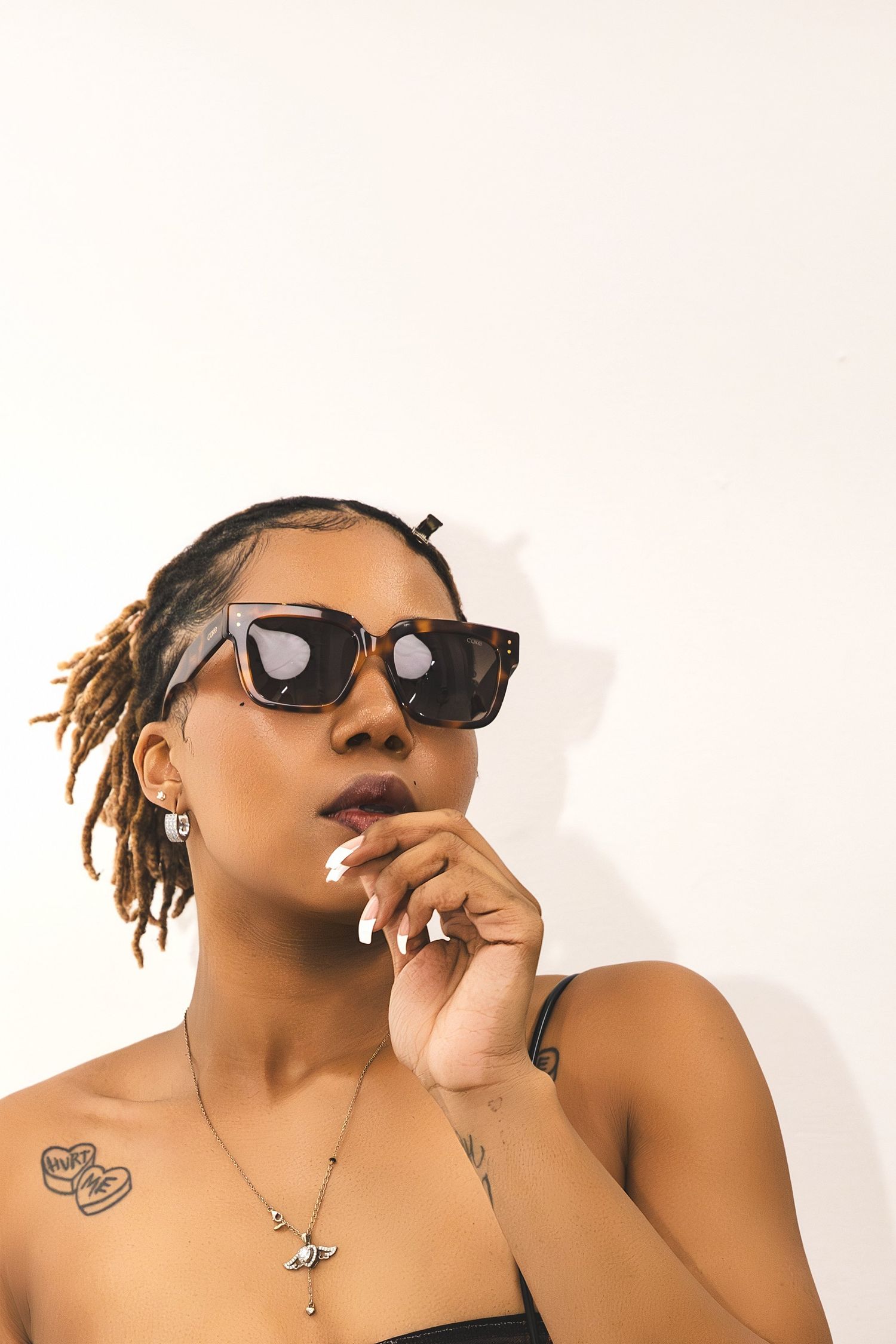 Young woman with locs wearing oversized tortoiseshell sunglasses, chest tattoos, and a necklace poses thoughtfully.