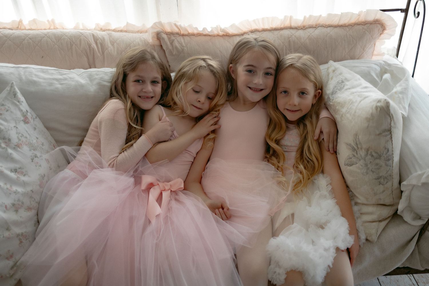 Group of young children in pink and white ballet outfits laughing and hugging together on a wooden floor.