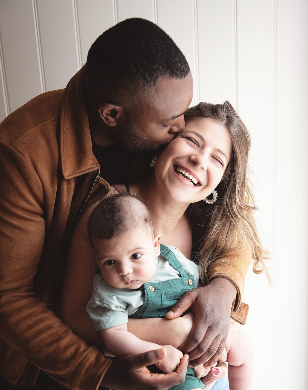 Happy interracial family portrait with father kissing mother's cheek while she smiles holding their newborn baby. During a family photoshoot
