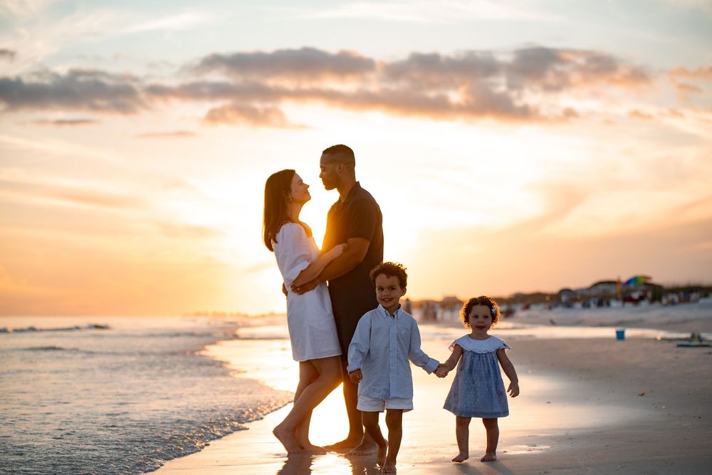 Family of four sharing a tender sunset moment on the beach, parents embracing while children hold hands by the ocean.