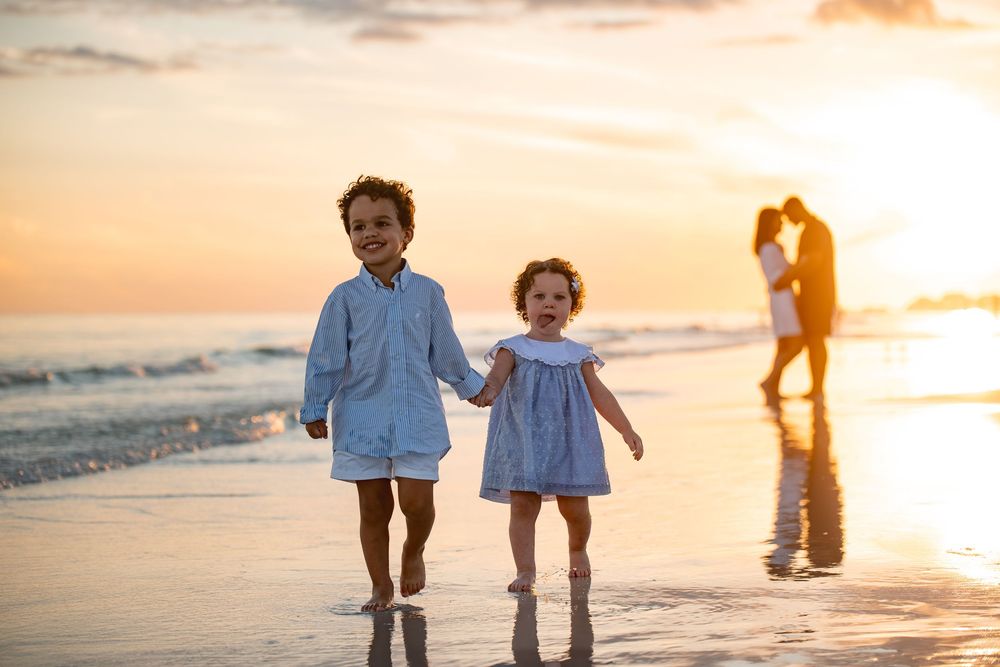 Two young children holding hands on a beach at sunset with their parents embracing in the background.