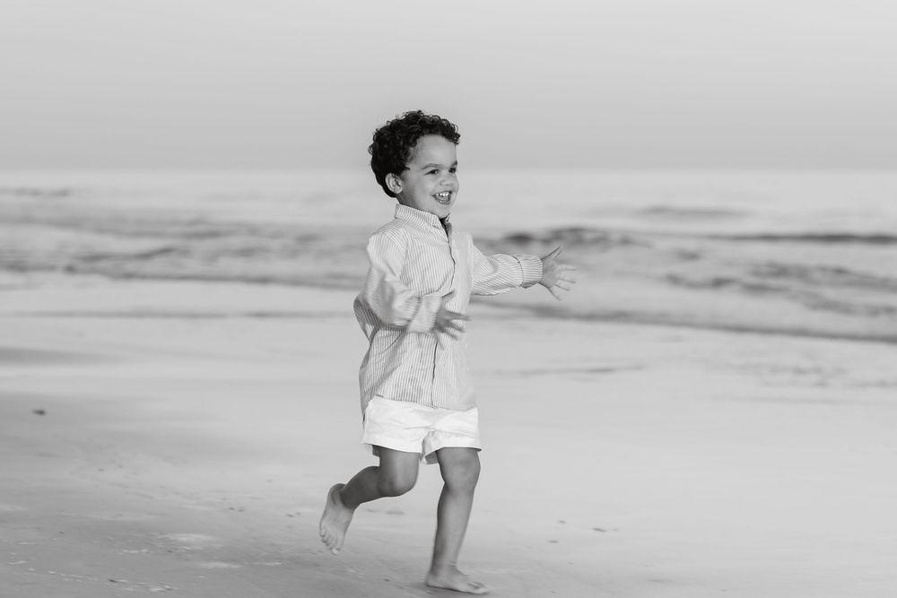 Happy young boy running barefoot along ocean shoreline in black and white photo.