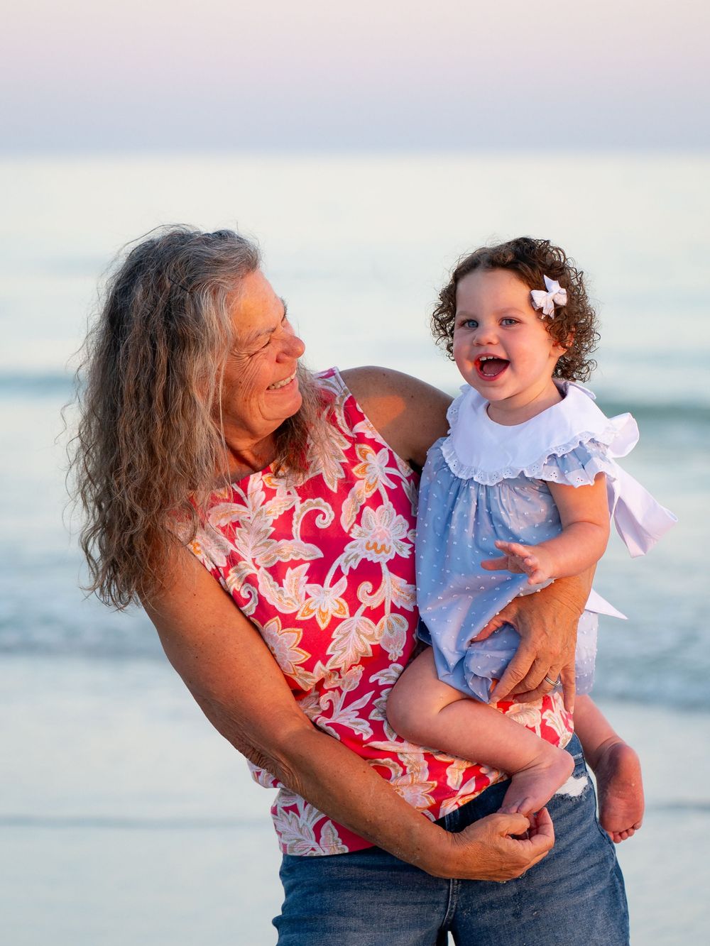 Grandmother joyfully holds a laughing toddler girl on a beautiful beach at golden hour.