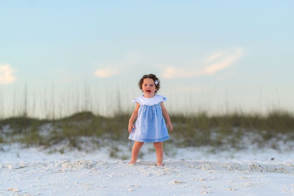 Joyful toddler girl in blue dress laughing on white sand beach with dunes and clear sky background.