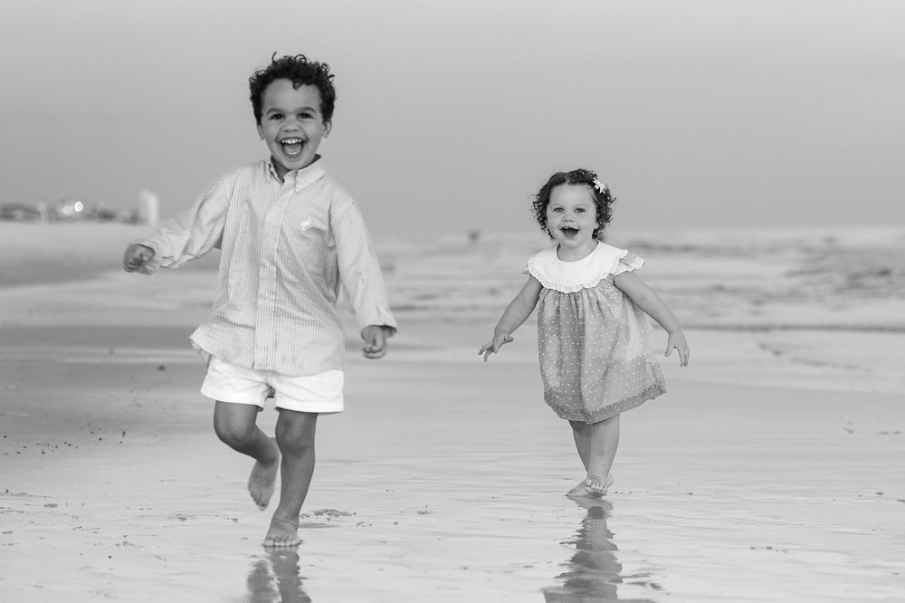 Two joyful children running and laughing on a sandy beach in black and white photography.