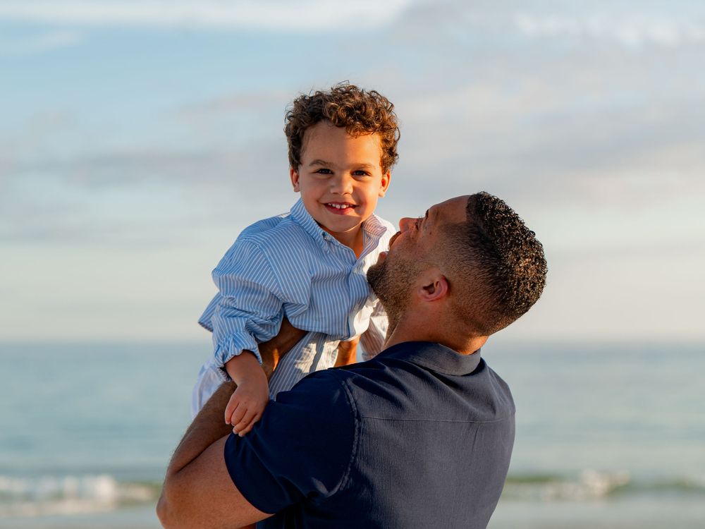 A father lovingly lifts his smiling toddler son on a sunny beach with ocean waves in the background.