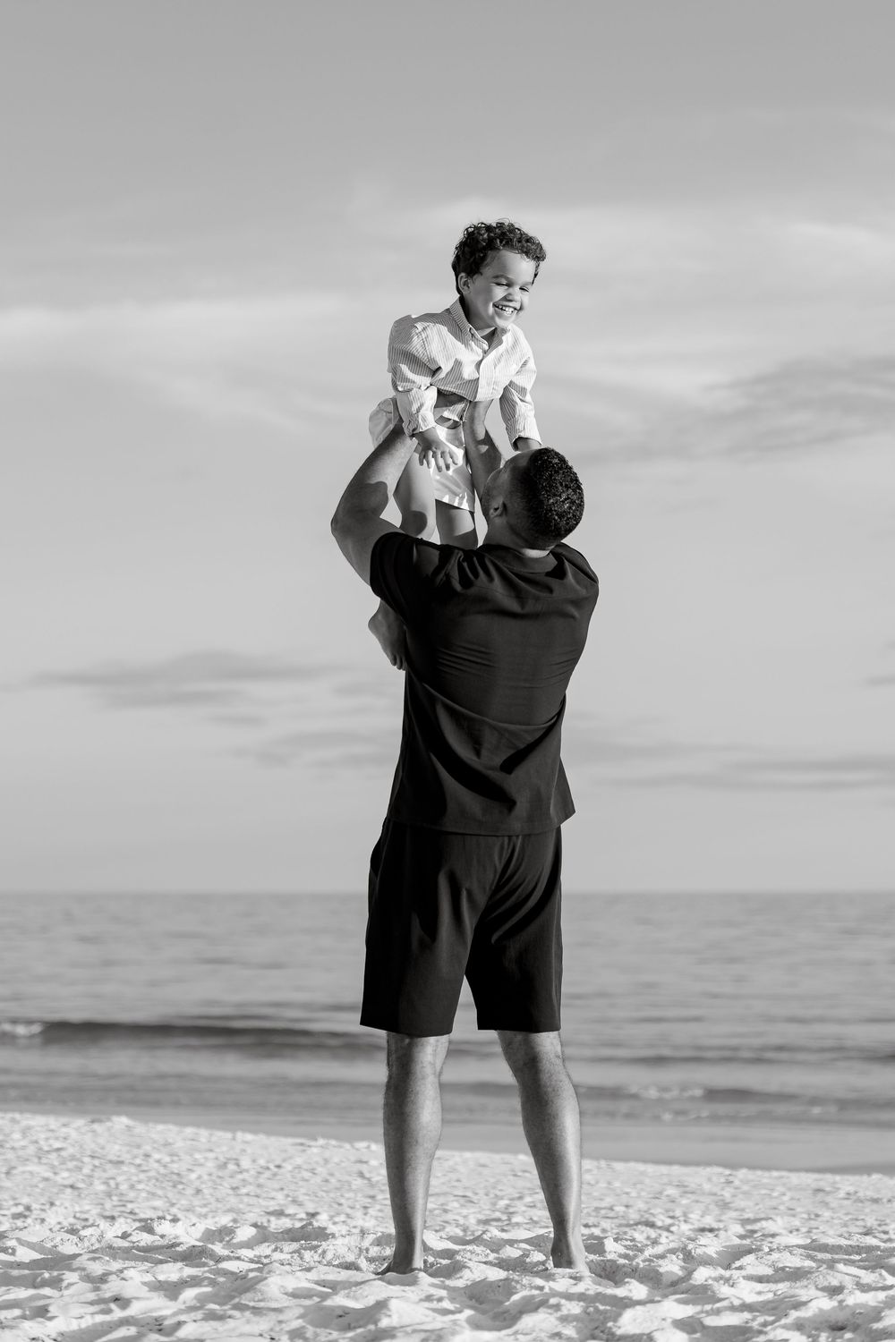 Father lifting laughing child overhead on sandy beach in black and white photo.