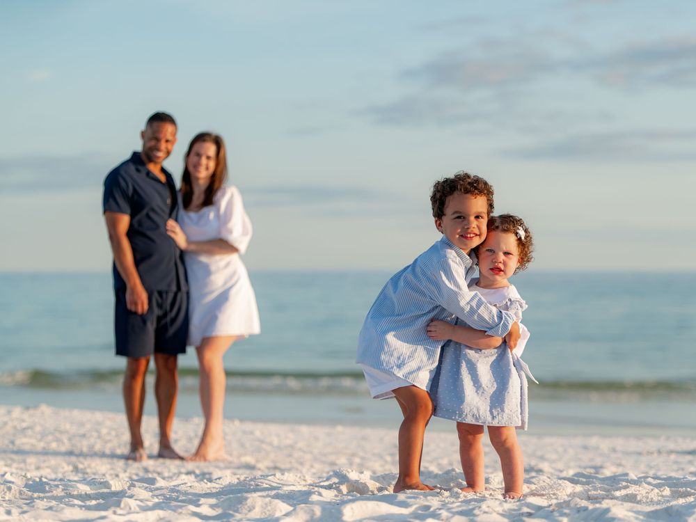 Family beach portrait with two young children hugging in foreground, parents standing together by ocean.
