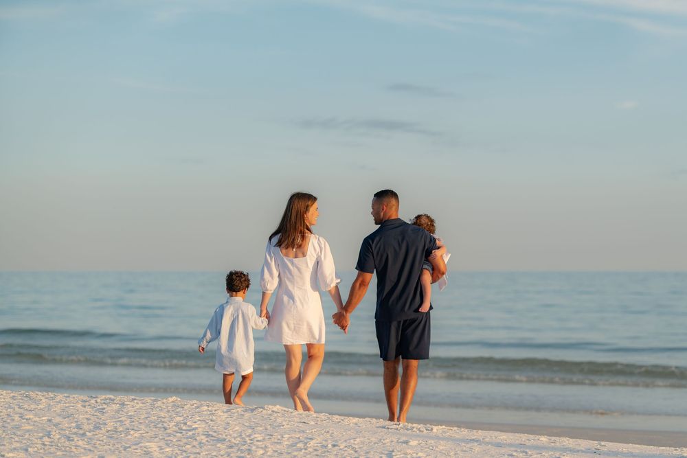 Family of four walking hand-in-hand along a serene white sand beach at sunset, viewed from behind.