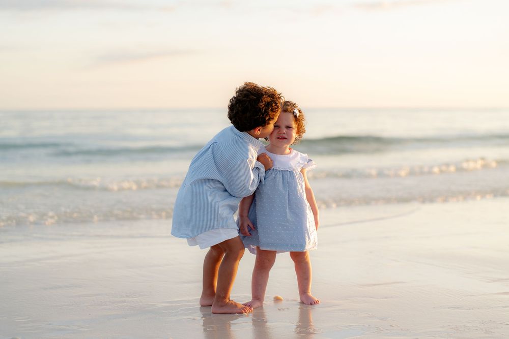 Two young children share a sweet moment on a sandy beach at sunset, waves gently rolling in behind them.