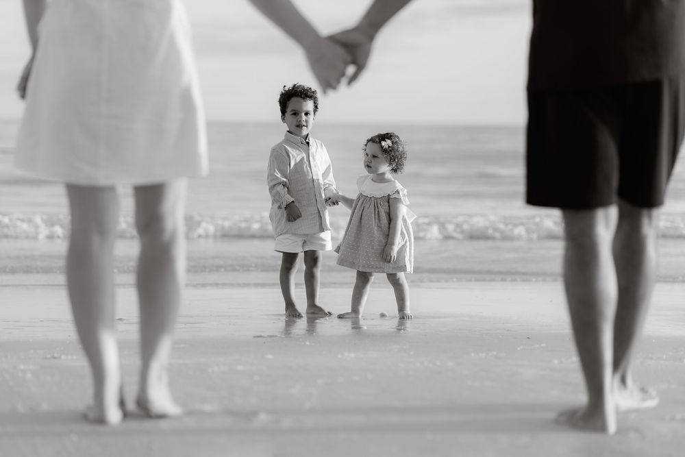Black and white beach photo of two young children holding hands between their parents by the ocean.