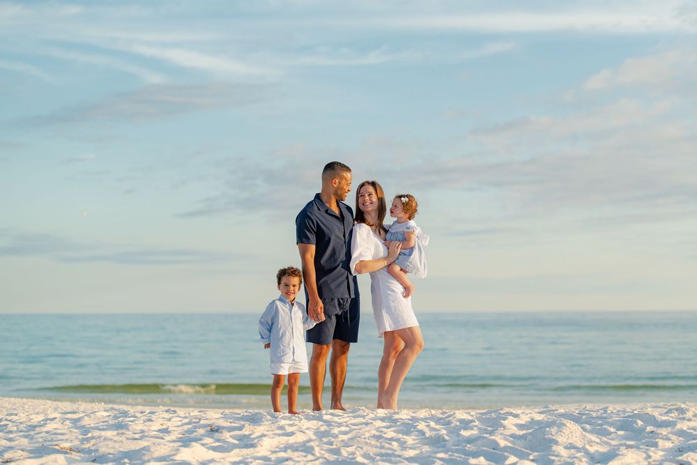 Family of four posing on a white sand beach with calm blue ocean and sky backdrop.