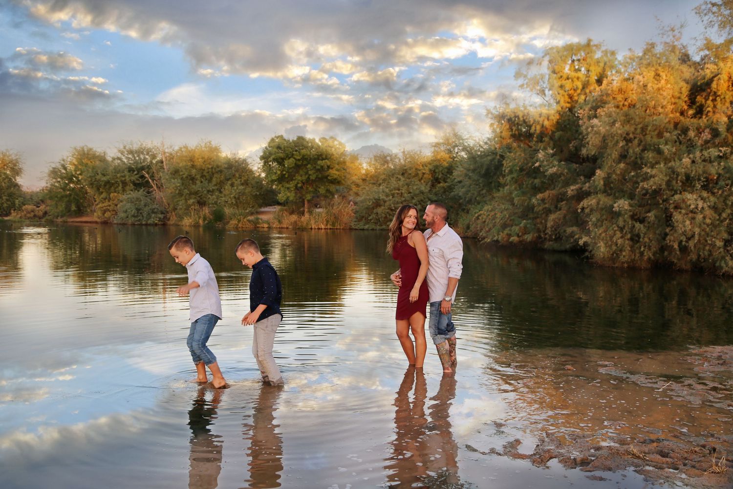 A family of four wades through shallow river water at golden hour, with trees reflecting on the surface and soft clouds above.