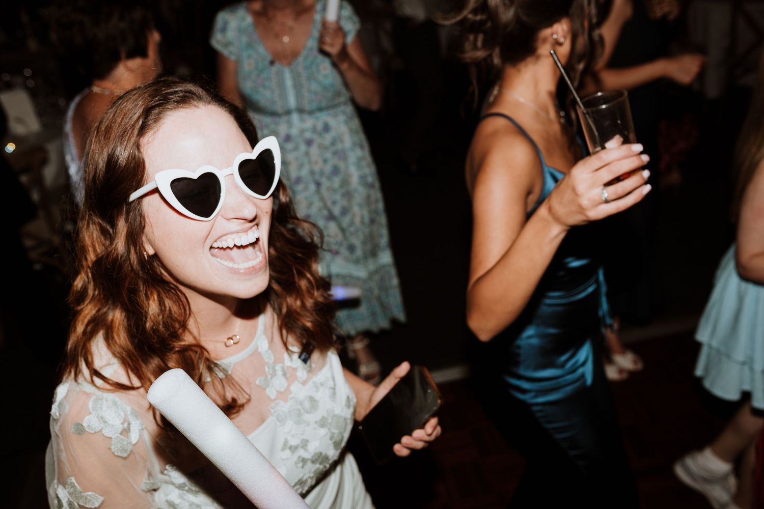 A joyful woman wearing heart-shaped white sunglasses dances at a party, holding a phone while others celebrate behind her.