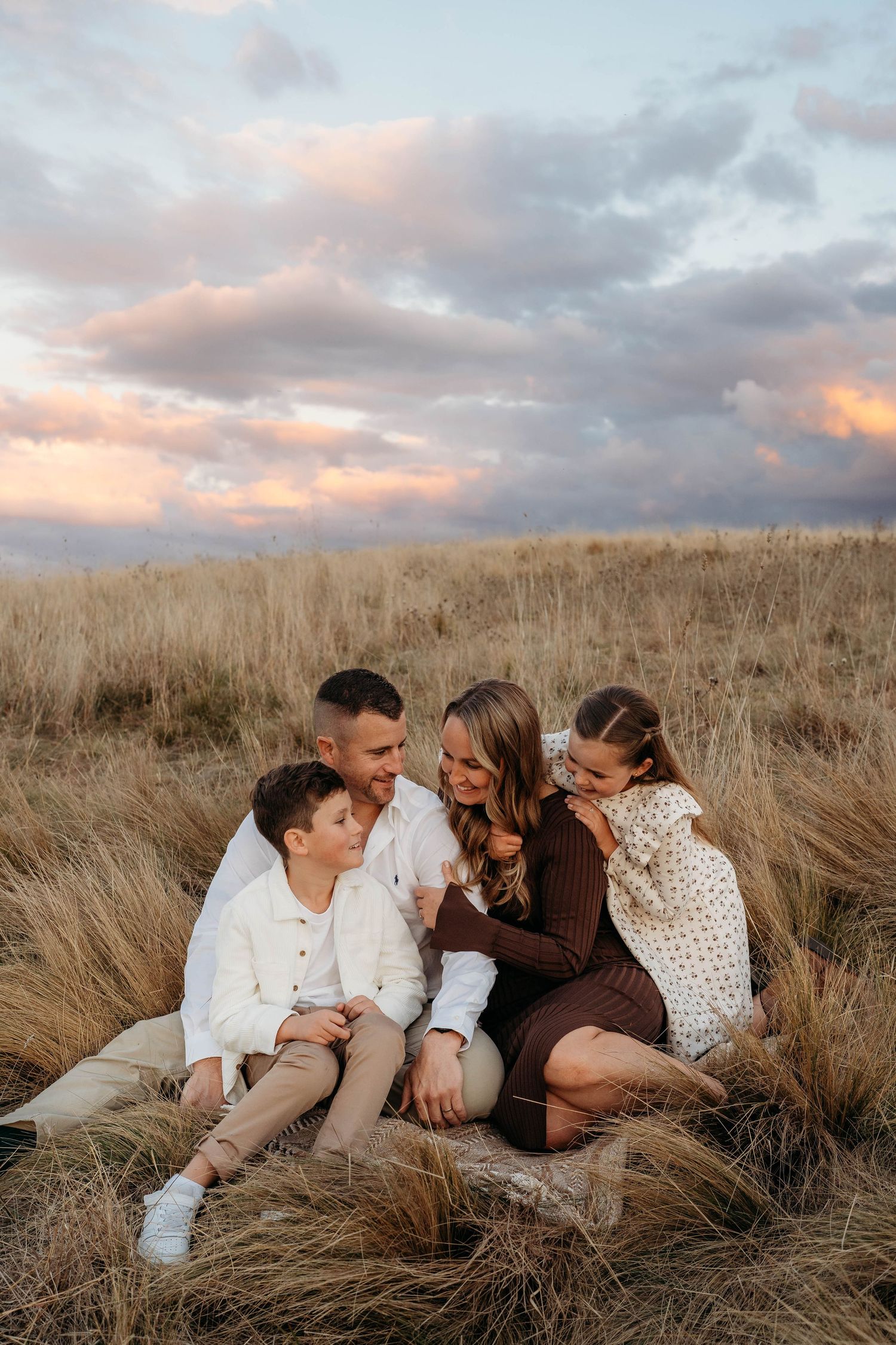 Happy family of four sitting together in golden grass field under colorful sunset cloudy sky.