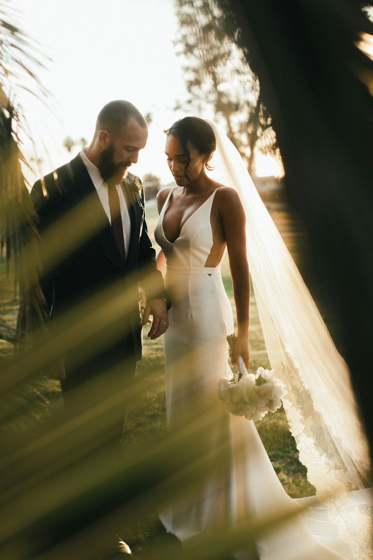 Bride in white gown with veil and groom in dark suit hold hands among palm trees in golden sunset light.