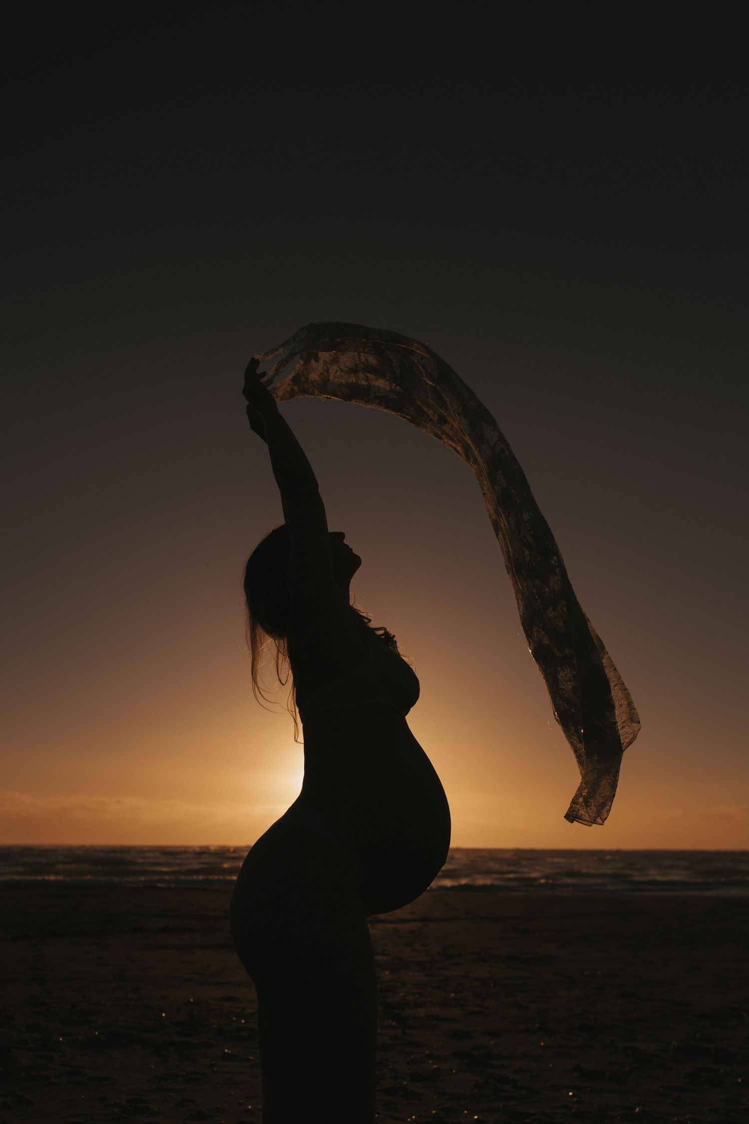 Pregnant woman standing in ocean waves at sunset, wearing jeans and a sweater on a sandy beach.