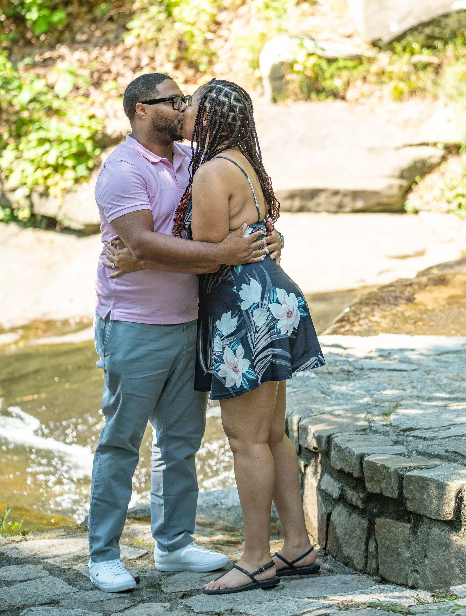 A couple shares a romantic kiss outdoors near a stone wall and stream, man in pink polo and woman in floral dress.