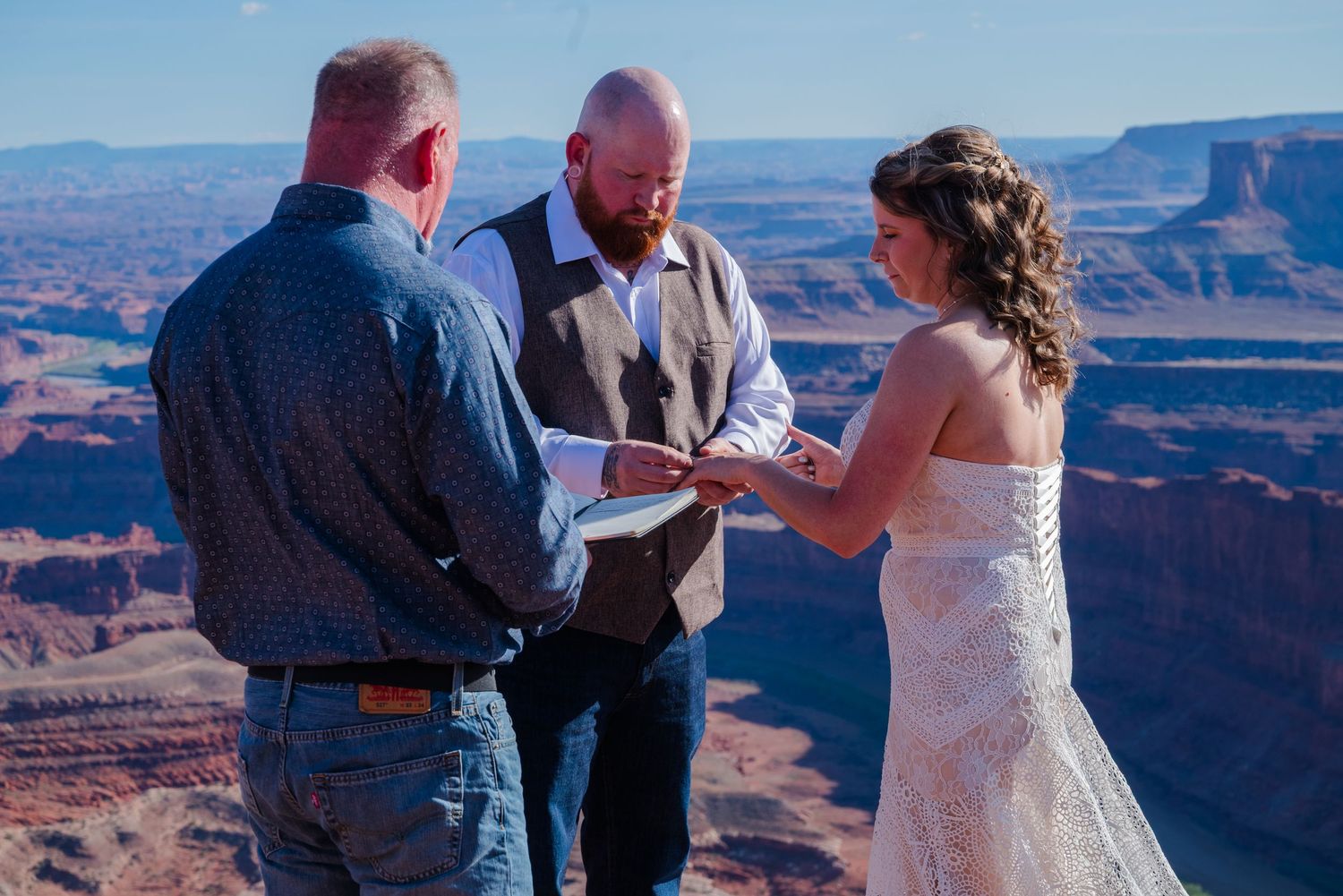 Couple exchanges rings during outdoor wedding ceremony at scenic red rock canyon overlook with stunning desert landscape backdrop.
