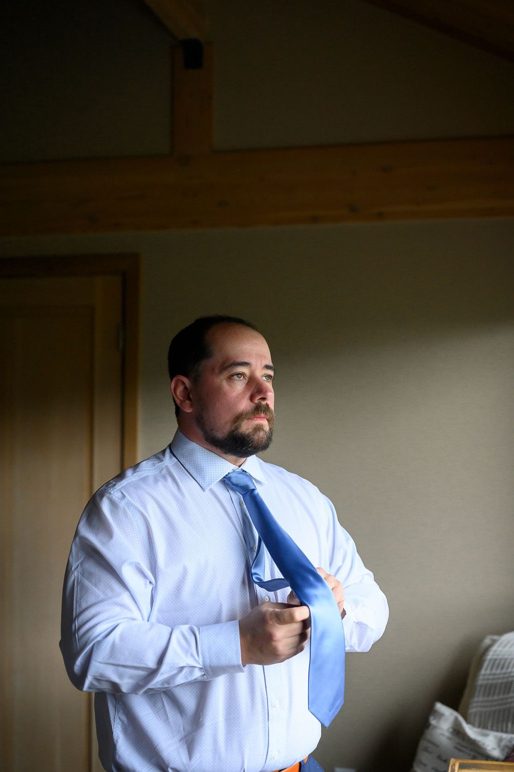 Groom adjusts his blue tie while getting ready indoors in soft natural window light.