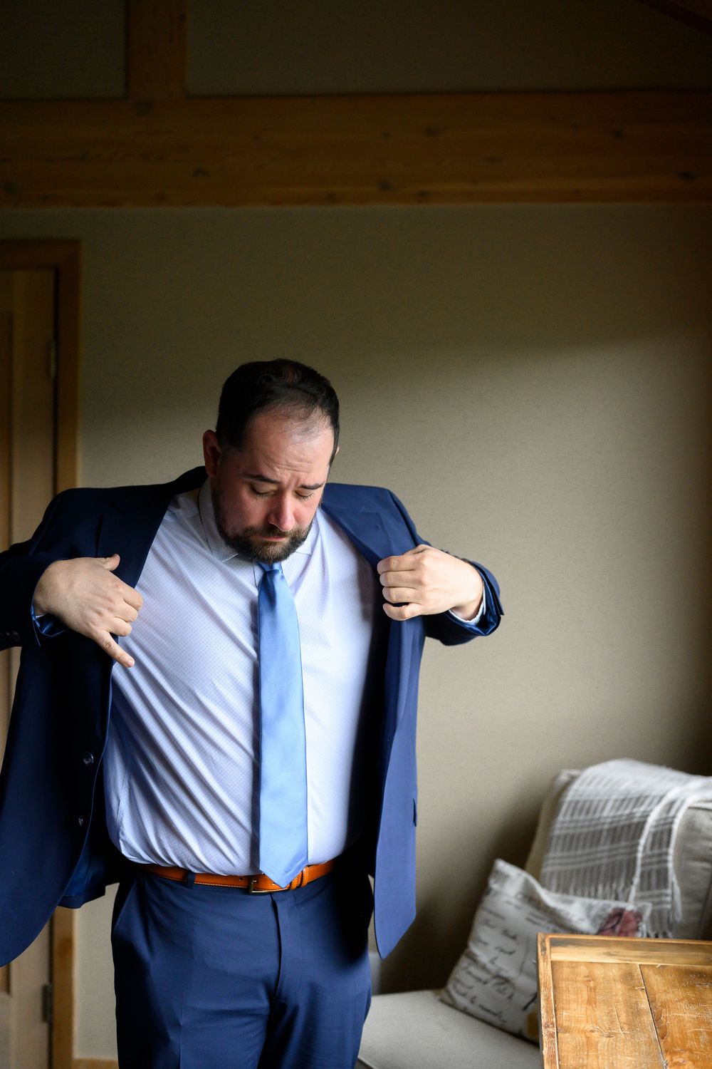 A man in a navy blue suit and light blue tie adjusts his jacket while getting dressed in a softly lit room.