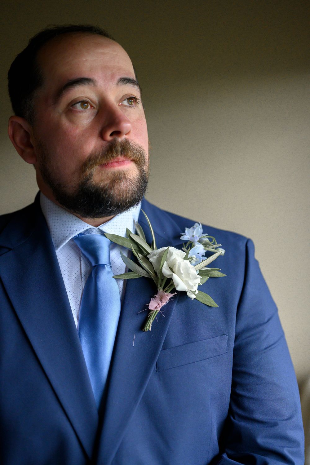 A bearded groom in a blue suit and tie with a floral boutonniere gazes upward thoughtfully.