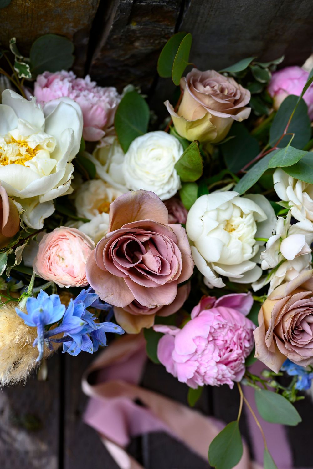Elegant floral bouquet featuring dusty pink roses, white peonies, blue flowers, and eucalyptus with a satin ribbon.