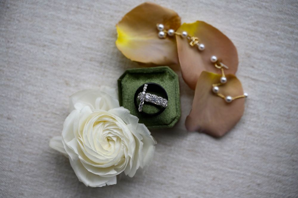 Diamond ring in green velvet box with white ranunculus, rose petals, and pearl bracelet on linen.