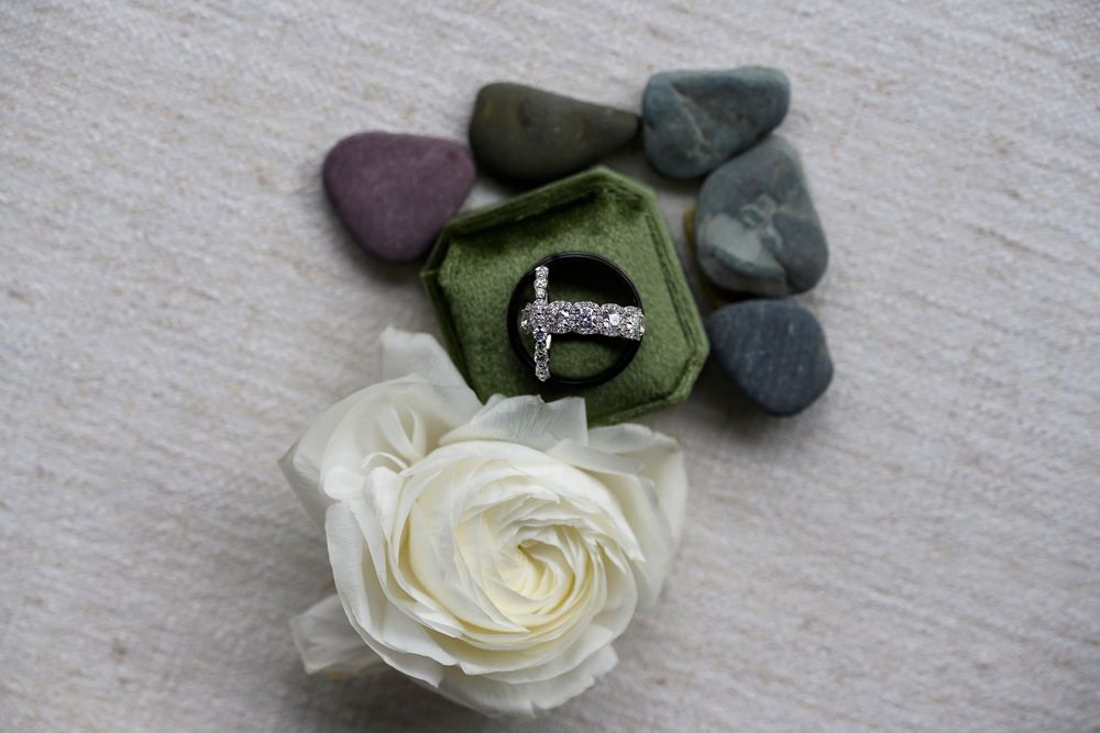 Silver cross ring displayed in a green velvet box, surrounded by smooth stones and a white rose on linen.
