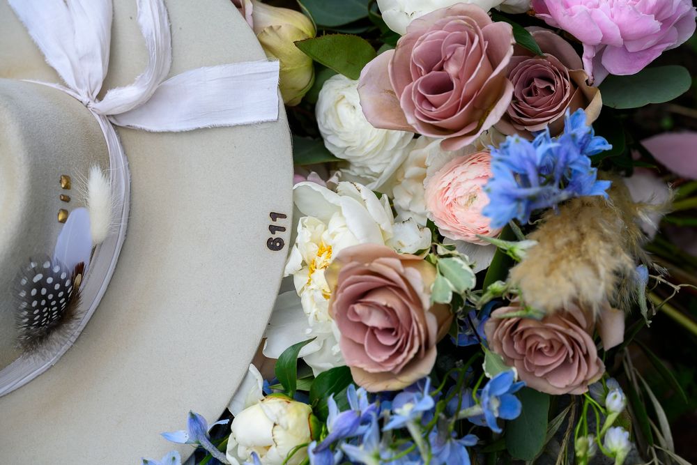 Elegant wedding table setting with dusty pink roses, white peonies, and blue delphinium flowers surrounding a numbered plate with a feather.