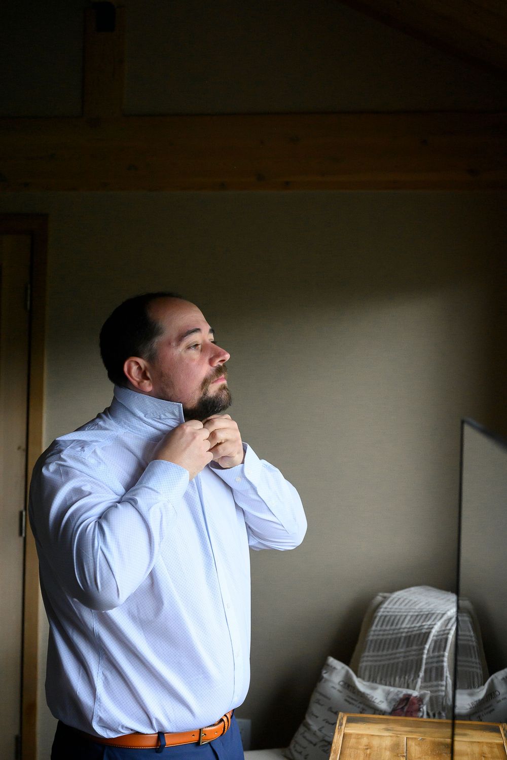 Man in white dress shirt adjusting his tie while getting dressed, standing in a dimly lit room.