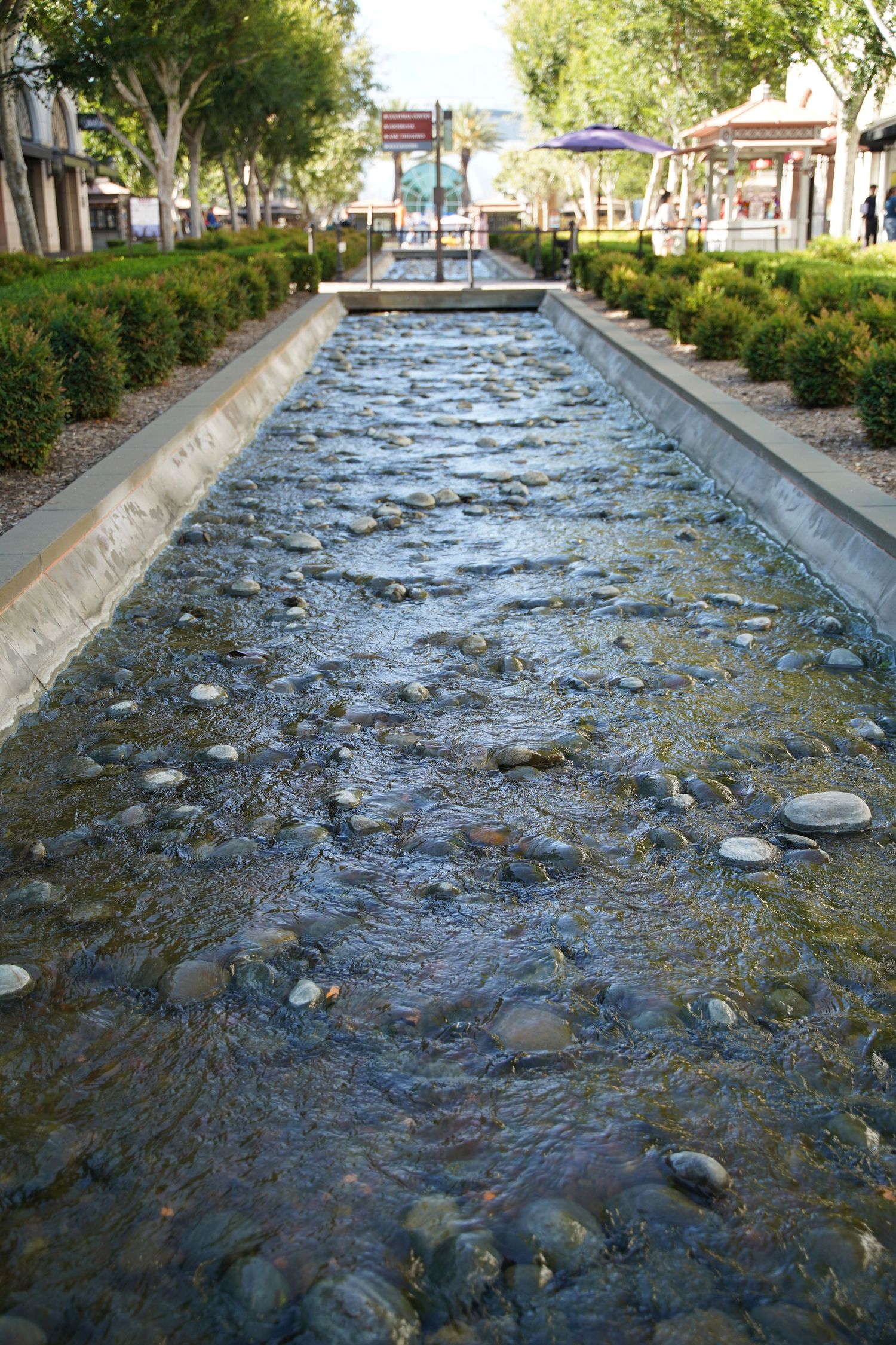 Shallow decorative water feature with river stones flows through a landscaped outdoor plaza surrounded by trees and shrubs.