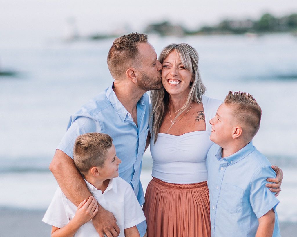 Happy family of four sharing a joyful moment on a beach, with parents and two young boys smiling and embracing.