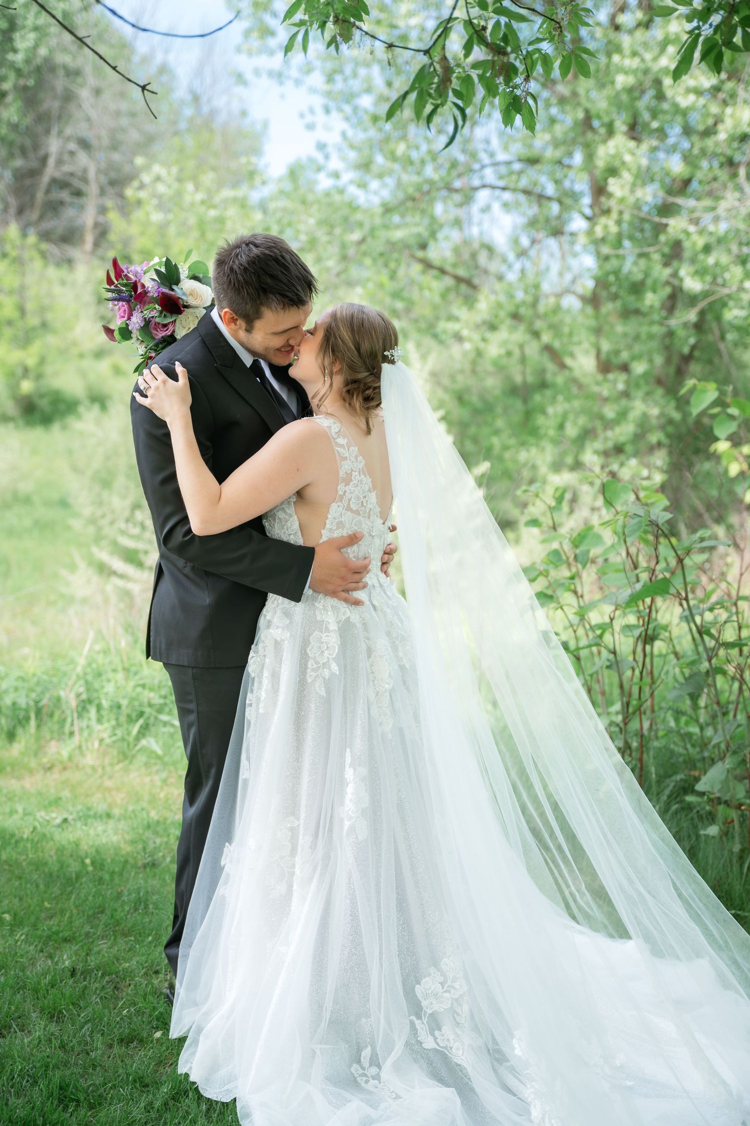 A bride and groom share a romantic kiss outdoors, surrounded by lush green trees, as her lace gown and veil flow elegantly.