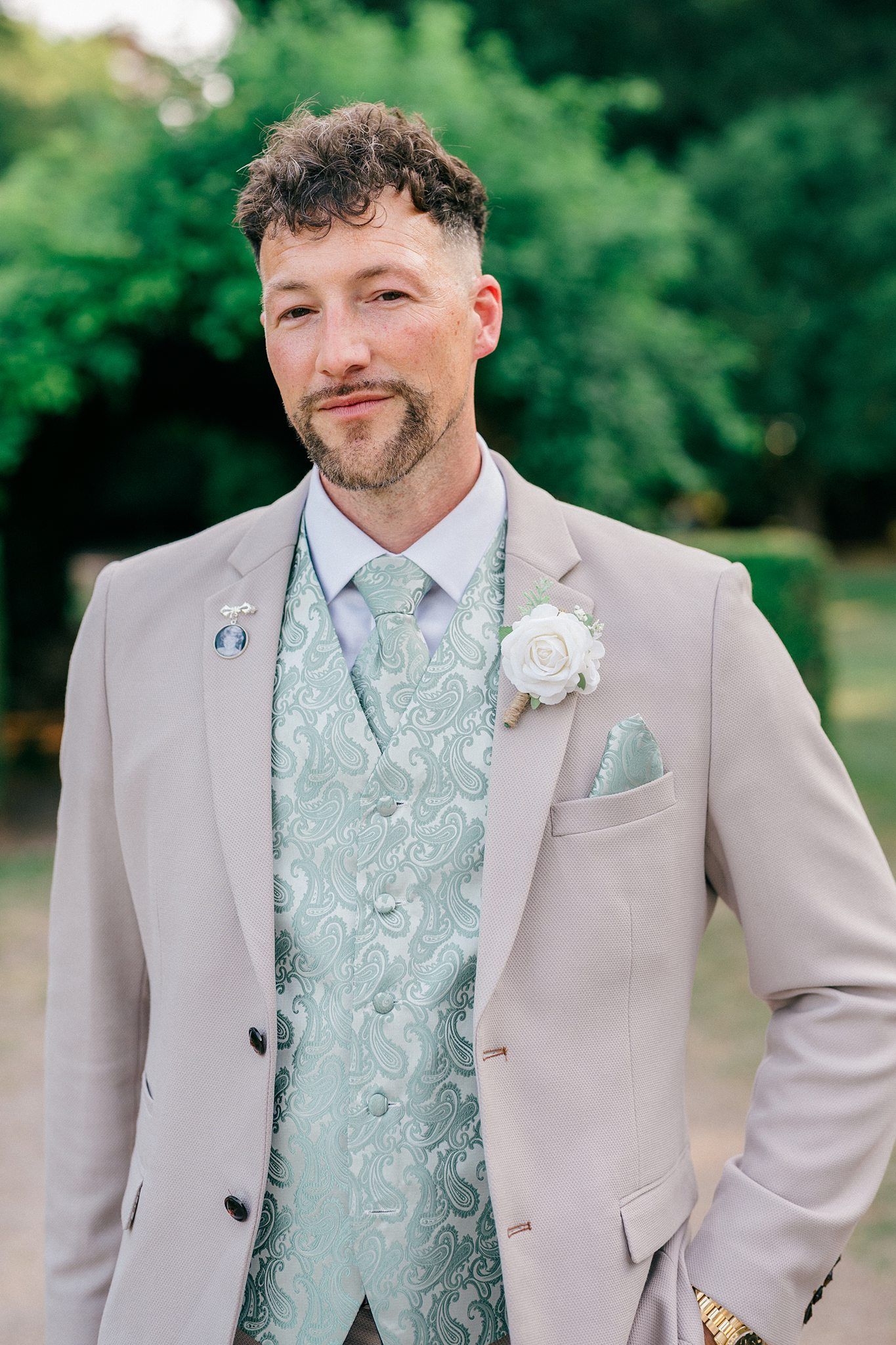 Groom in a light beige suit with mint green paisley waistcoat and tie, white rose boutonniere, posing outdoors.