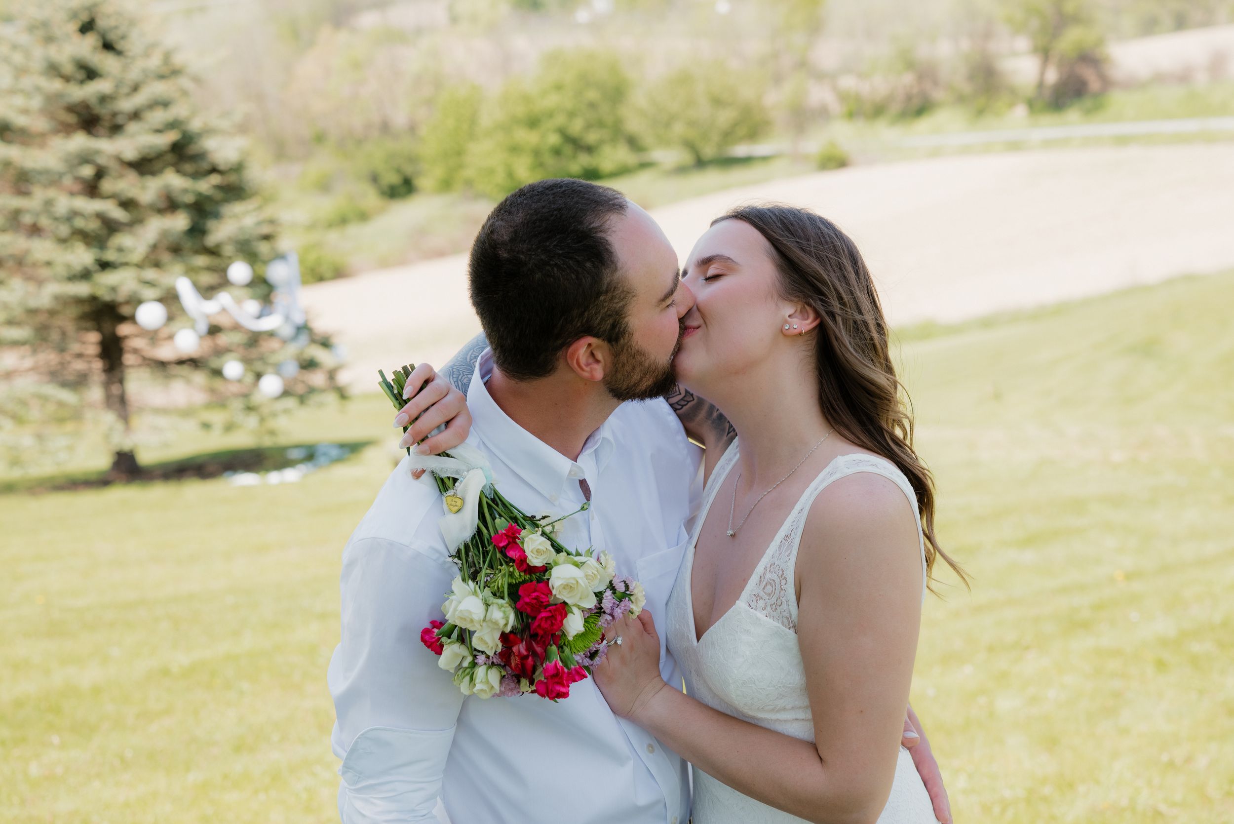 A newlywed couple shares a romantic kiss outdoors, the groom in white holding a red and white bouquet.